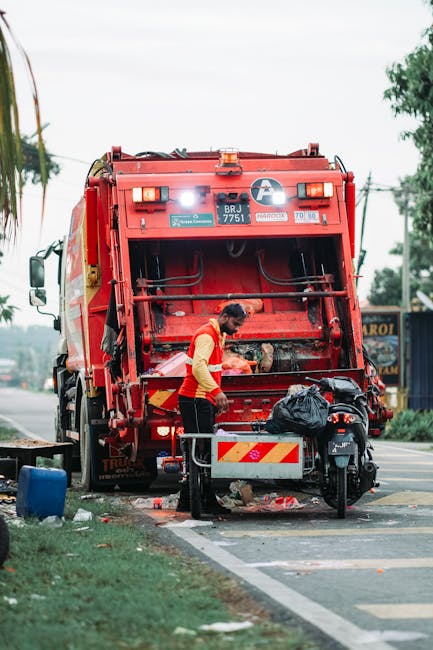 A worker in a yellow and red uniform is standing at the rear of a large red rubbish collection truck, which is parked on the side of a street. The truck's rear door is open, revealing the compaction mechanism and emptying area. The worker appears to be handling rubbish or equipment, with a black trash bag placed on a small metal platform attached to the back of the truck. Surrounding the scene, there are various objects including a black motorbike parked nearby, some scattered waste on the pavement, and a blue container on the grass verge. The background features trees, utility poles, and a building with signage, indicating an urban or suburban area. The environment is daylight with an overcast sky, and the scene reflects on-site waste collection activities relevant to private rubbish removal and alternative disposal methods, such as quick or independent collection services provided by companies like Rubbish Removal Barnet.