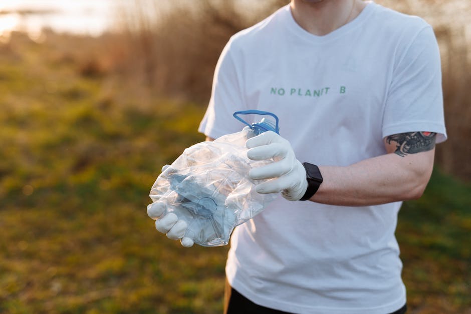 A person in a white T-shirt with the text 'NO PLANT B' printed in green is standing outdoors on a grassy area during daylight, holding a transparent plastic bag filled with crumpled white rubbish or waste material. The individual is wearing white gloves and a black wristwatch on their left wrist, and has a tattoo on their right forearm. The background features a natural environment with blurred trees and foliage, illuminated by warm, diffused sunlight, suggesting an informal outdoor waste collection or clean-up activity that relates to private rubbish handling or independent waste disposal. The scene emphasizes responsible waste management practices in a natural setting, supporting rubbish removal services like those provided by Rubbish Removal Barnet.