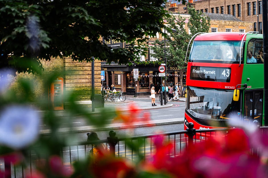 A double-decker bus with a red and green livery traveling along a city street in Barnet, with the route number 43 and destination 'Friern Barnet' displayed on the front. The bus is positioned to the right of the image, partially obscured by vibrant pink, red, and white flowers in the foreground and leafy green branches at the top. Behind the bus, there is a busy intersection with pedestrians crossing the road and standing on the sidewalk, some engaging with bicycles parked nearby. On the opposite side of the street, a traditional pub with dark wooden signage and multiple windows is visible, along with other old brick buildings contributing to a historic urban atmosphere. The scene is illuminated with natural daylight, casting soft shadows and enhancing the warm, lively mood typical of public transport and street life in Barnet, reflecting the area's diverse and active community. This visual context subtly aligns with services like independent rubbish collection or private waste management, commonly seen in urban areas with busy streets and commercial establishments, as provided by Rubbish Removal Barnet.