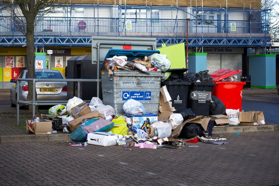 A large collection of overflowing rubbish bins and scattered waste bags, including black, white, and red trash bags, cardboard boxes, and paper waste, is piled on a paved area next to a sidewalk in an urban setting. The waste is situated in front of two black bins and a grey recycling container designated for mixed paper and cardboard, with some lids open and contents spilling over. Behind the waste, a silver car is parked parallel to a metal railing, and in the background, there are storefronts with signage, a blue construction fence, and a building under renovation. The scene appears to illustrate an unmanaged waste accumulation outside commercial premises, highlighting the importance of proper rubbish management, potentially related to private or alternative waste disposal services like those offered by Rubbish Removal Barnet, in contrast to council-run collections.