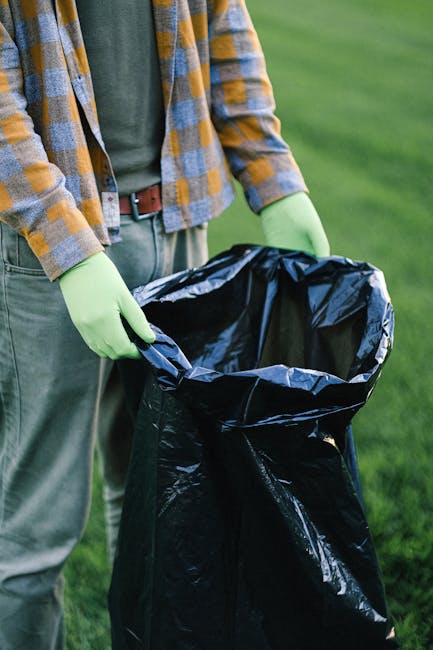 A person wearing a plaid shirt in shades of yellow and grey, along with green gloves, is holding open a large black rubbish bag made of shiny plastic material. The bag is partially filled with waste and is positioned outdoors on a grassy area, likely a park or garden, with a blurred green background. The individual’s torso, arms, and hands are visible, but not their face. The scene suggests a process of waste collection or rubbish removal, possibly as part of a private clearance service by Rubbish Removal Barnet, and emphasizes careful handling of waste materials. The environment appears well-lit, with natural daylight illuminating the scene, creating a clear, professional view relevant to rubbish collection and disposal activities.