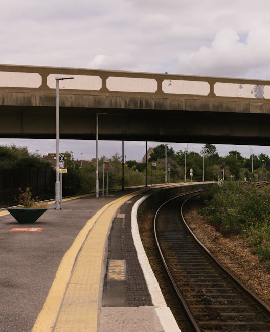 A railway station platform situated underneath a concrete overpass bridge, with an elevated road running above. The platform features a yellow safety line along the edge, a tactile paving strip, and a single lamppost placed near the platform's middle. The surface of the platform appears clean and smooth, with some greenery growing alongside the tracks and bushes lining the perimeter. The railway tracks curve gently to the right, extending into the distance, with dark, metallic rails and wooden sleepers visible beneath. The sky overhead is partly cloudy, creating diffuse natural light that highlights the textures of the concrete bridge, the railway surface, and the surrounding vegetation. The scene reflects a typical urban or suburban setting where private or independent waste removal services, such as those offered by Rubbish Removal Barnet, might operate in the vicinity of public transportation infrastructure, contributing to efficient local waste management and alternative rubbish collection options.